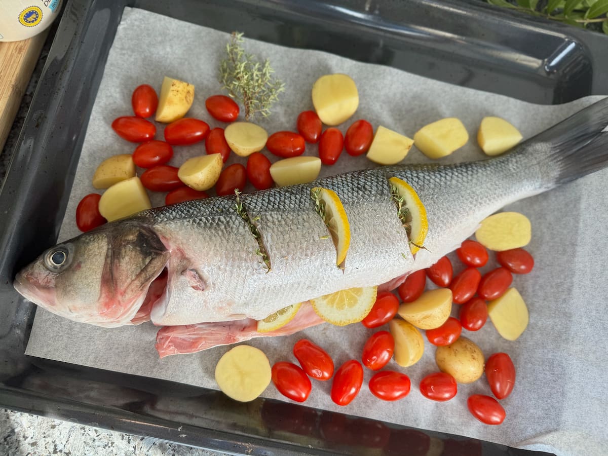 whole fish on a roasting tin with baking parchment and skin scored, stuffed with lemon, herbs and surrounded with potatoes and cherry tomatoes