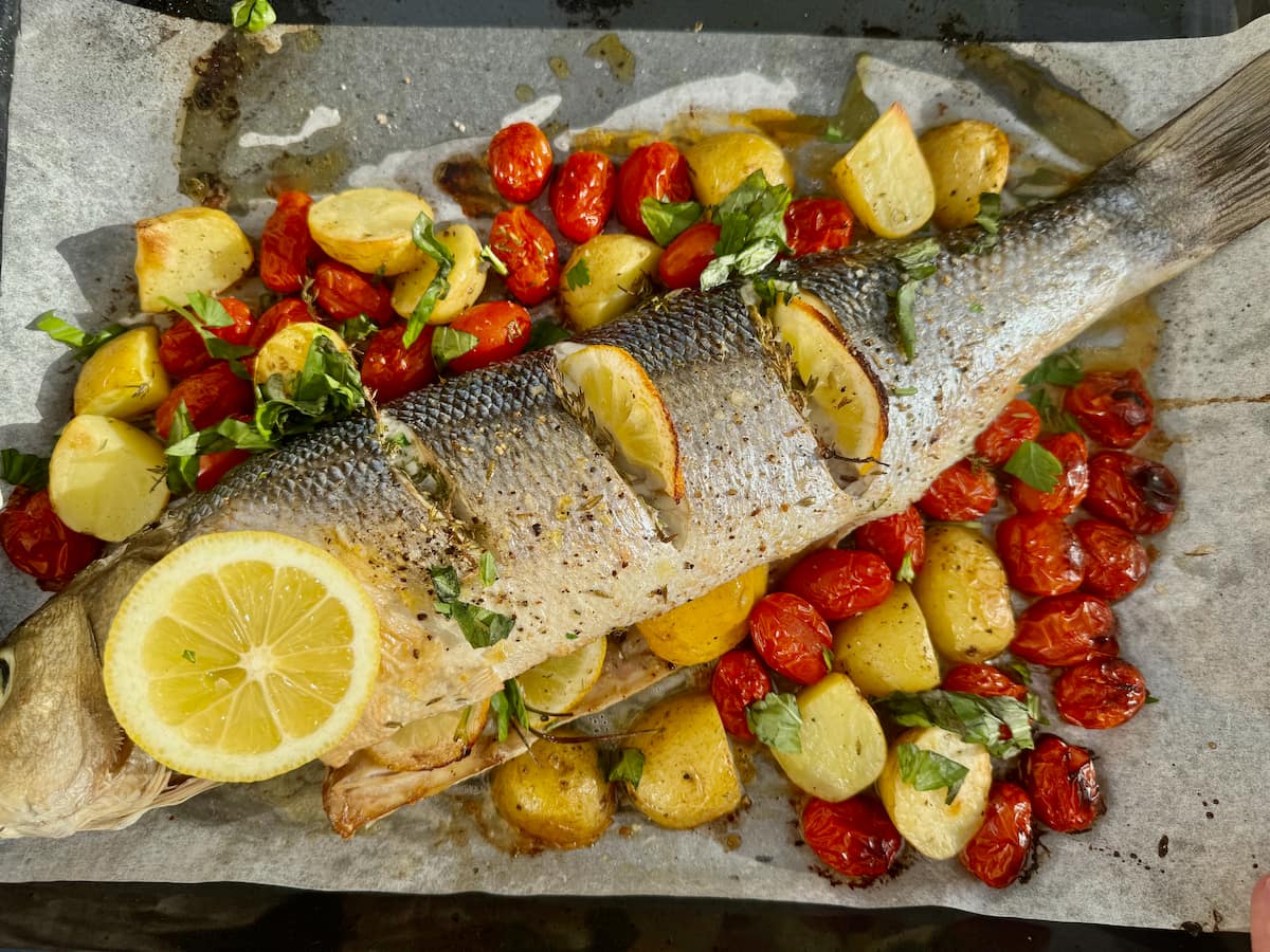 whole sea bass fish baked in a roasting dish with cherry tomatoes, potatoes, herbs and lemon