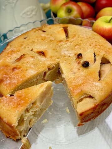 large apple cake showing moist inside with a slice removed, topped with a shiny glaze on a plate next to apples